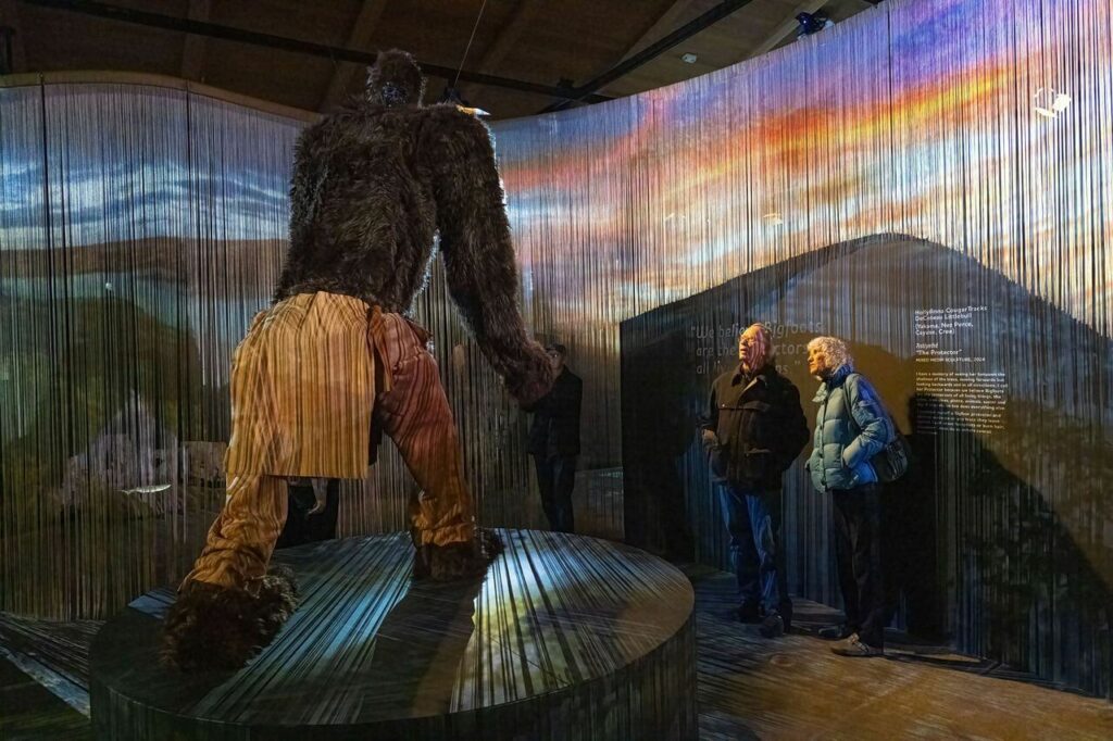 Replica of sasquatch in tan pants with museum spectators admiring its splendor, set against an exhibit of strings that form to make a visual of a sunset.