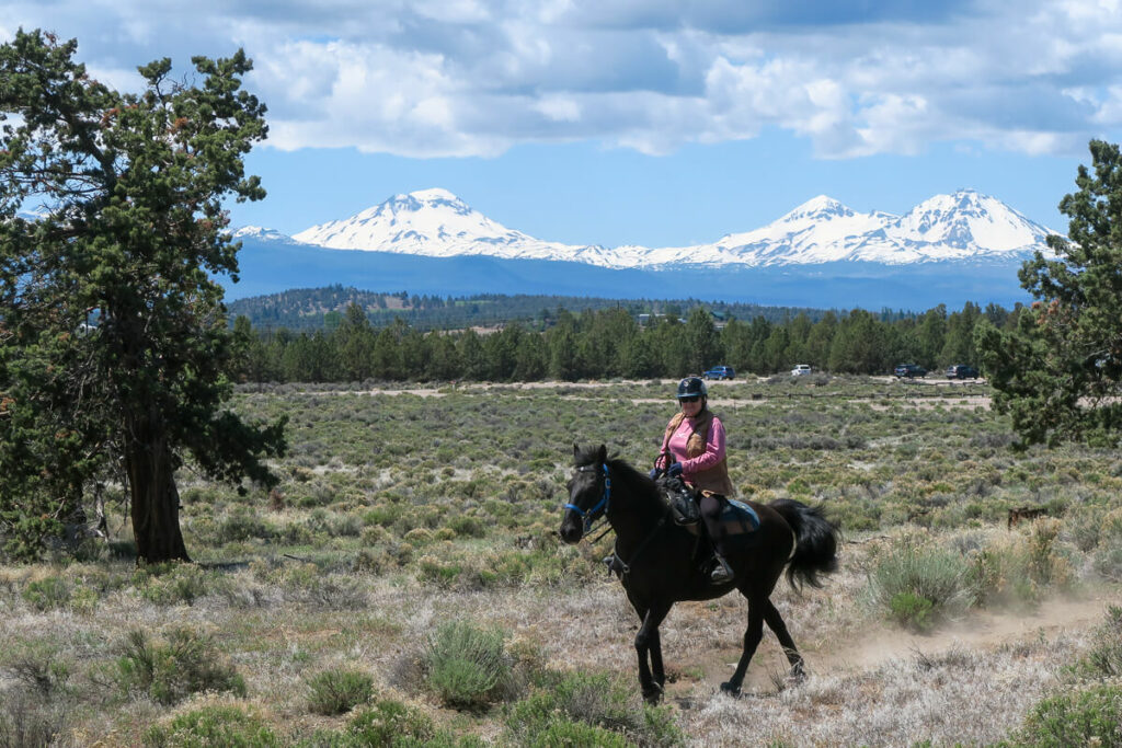Two women in pink and blue tops riding horses in front of the Three Sisters mountain peaks.
