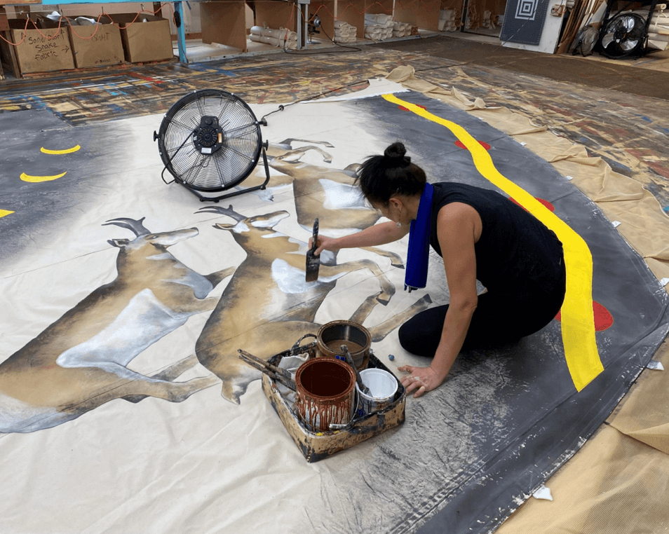 Woman sitting on the ground painting antelope on the canvas of a teepee.